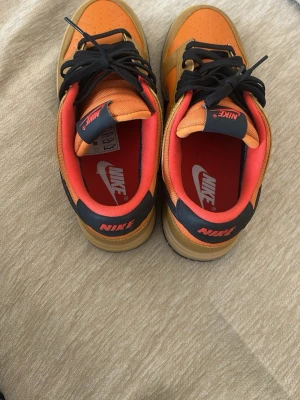 Nike Dunk Low orange och beige - Säljer ett par Nike Dunk Low sneakers i orange och beige med svarta detaljer och svart swoosh. Skorna har svart snörning, röd insida och klassisk Nike-logga på hälen. Ovandelen är i läder och mesh för en schysst look.