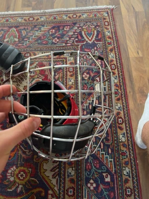 Ishockeygaller i krom - Säljer ett klassiskt ishockeygaller i silverfärgad metall med svart vaddering vid hakan. Gallret passar till hjälm och skyddar ansiktet under spel. Perfekt för dig som vill ha extra säkerhet på isen.