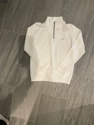 Vit half-zip sweatshirt från Moncler - Snygg vit sweatshirt från Moncler med half-zip framtill och hög krage. Tröjan har ett diskret Moncler-märke på bröstet och är gjord i mjukt material. Långa ärmar och ribbade muddar ger en clean och stilren look. Perfekt för dig som gillar exklusiva basics.