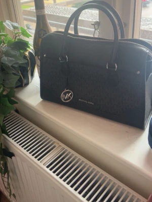 Svart handväska från Michael Kors - Snygg svart handväska från Michael Kors med klassiskt MK-mönster och silverfärgade detaljer. Väskan har dubbla handtag, dragkedja upptill och en rund MK-berlock framtill. Tillverkad i tåligt konstläder med stilren design.