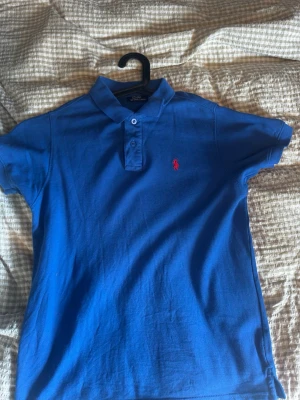 Blå pikétröja från Ralph Lauren - Klassisk blå pikétröja från Ralph Lauren med korta ärmar och krage. Tröjan har två knappar framtill och den ikoniska röda loggan broderad på bröstet. Tillverkad i mjuk bomull som känns skön mot huden. Perfekt till jeans eller shorts.