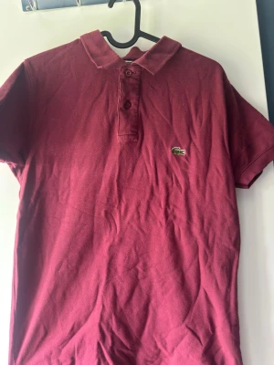 Vinröd pikétröja från Lacoste - Klassisk vinröd pikétröja från Lacoste med korta ärmar och krage. Tröjan har knappslå framtill och den ikoniska gröna krokodilloggan på bröstet. Tillverkad i mjuk bomull som känns skön mot huden. Perfekt för en avslappnad men ändå stilren look.