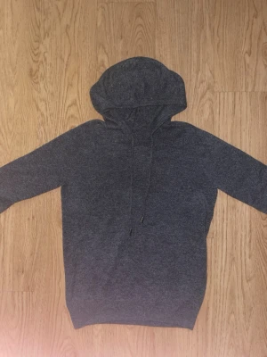 Mörkgrå kashmir hoodie med dragsko - En enkel och stilren mörkgrå hoodie med huva och dragsko. Hoodien har långa ärmar och ribbade muddar vid ärmslut och nederkant. Perfekt för en avslappnad och bekväm look. Materialet känns mjukt och stretchigt, troligen bomullsblandning.