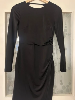 Svart långärmad klänning med drapering - Stilren svart klänning med lång ärm och rund halsringning. Klänningen har en snygg drapering på sidan och är tillverkad i ett mjukt, stretchigt material. Passar perfekt för dig som gillar en enkel men elegant look.