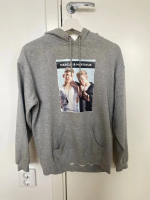 Grå Marcus & Martinus hoodie - Säljer en grå hoodie med tryck av Marcus & Martinus på bröstet. Hoodien har huva med dragsko och en stor magficka framtill. Tillverkad i mjukt sweatshirtmaterial och har ribbade muddar vid ärmar och nederkant. Perfekt för dig som gillar snygga och bekväma plagg.