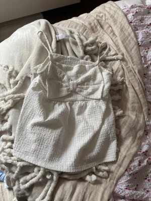 Vit linnetopp med rosett - Söt vit linnetopp med tunna axelband och en stor rosett framtill. Toppen har en lätt strukturerad yta och är croppad i modellen. Perfekt för varma dagar och enkel att styla med jeans eller kjol.