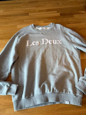 Grå sweatshirt från Les Deux - Snygg grå sweatshirt från Les Deux med vit broderad logga på bröstet. Tröjan har rund halsringning, ribbade muddar vid ärmslut och nederkant samt en liten tygpatch med logga nertill. Perfekt för en avslappnad och stilren look.