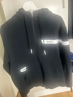 Svart zip-hoodie från BOSS - Snygg svart hoodie med dragkedja från BOSS. Hoodien har vita detaljer, justerbar huva med snören och en stor ficka framtill. På bröstet finns en vit rand med BOSS-logga. Perfekt för en avslappnad och trendig stil.
