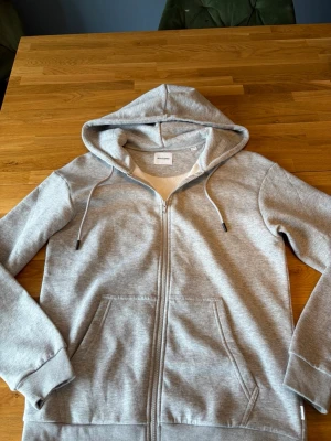 Grå zip hoodie från Jack & Jones - Snygg gråmelerad hoodie från Jack & Jones med dragkedja framtill, två stora fickor och justerbar huva med snören. Tillverkad i mjuk bomullsblandning och har ribbade muddar vid ärmar och nederkant. Perfekt för en avslappnad och stilren look.aldrig använd