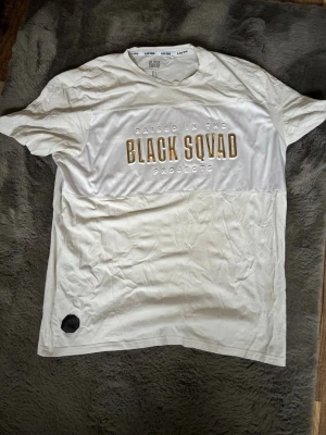 Vit T-shirt från Black Squad XL - Vit T-shirt från Black Squad med trycket 'RAISED IN THE BLACK SQUAD PROJECTS' i guld och vitt på bröstet. Regular fit med korta ärmar och rund hals. Tillverkad i mjuk bomull, perfekt för en clean och stilren look.