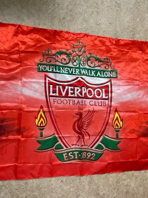 Liverpool FC supporterflagga - Stor röd supporterflagga med Liverpool Football Club-logga, texten 'You'll Never Walk Alone' och detaljer i grönt, vitt och gult. Perfekt för fans som vill visa sitt stöd för laget. Tillverkad i tunt tygmaterial.