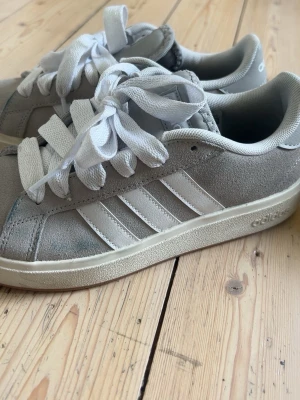 Grå Adidas sneakers i mocka - Snygga grå sneakers från Adidas med klassiska vita ränder på sidan. Skorna är låga och har ovandel i mocka, vita skosnören och en vit sula. Perfekta för dig som gillar stilrena och sportiga skor med ikonisk design.