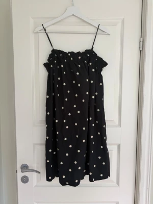 Svart prickig kort klänning H&M - Säljer en svart kort klänning från H&M med vita prickar och tunna axelband. Klänningen har en rynkad överdel och är perfekt för varma dagar. Lätt och luftig modell som ger en avslappnad känsla.
