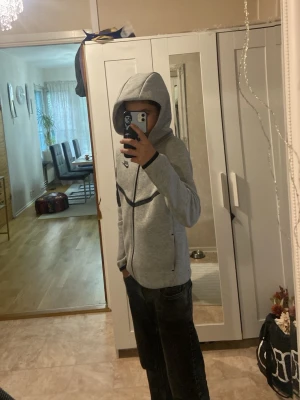 Grå Nike hoodie med dragkedja - Snygg grå hoodie från Nike med hel dragkedja och svarta detaljer längs dragkedjan, ärmslut och bröstet. Hoodien har en huva och en extra ficka med dragkedja på ena ärmen. Perfekt för en sportig och avslappnad stil.