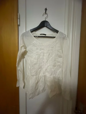 Vit blus med broderi och volang - Söt vit blus från H&M med broderade detaljer framtill och volang nedtill. Blusen har rund halsringning och långa ärmar. Tillverkad i ett lätt och luftigt tyg som ger en romantisk känsla.