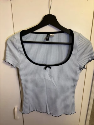 Ljusblå ribbad topp från H&M Divided - Supersöt ljusblå ribbad topp från H&M Divided med svarta detaljer. Toppen har korta ärmar, en djup rundad urringning med svart kant och en liten rosett framtill. 