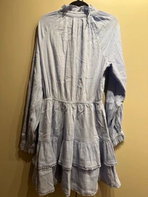 Ljusblå volangklänning med knyt - Supersöt ljusblå kortklänning med volanger och knyt i halsen. Klänningen har långa ärmar, knappar framtill och en luftig passform. Perfekt för sommarens alla tillfällen och tillverkad i mjuk bomull.