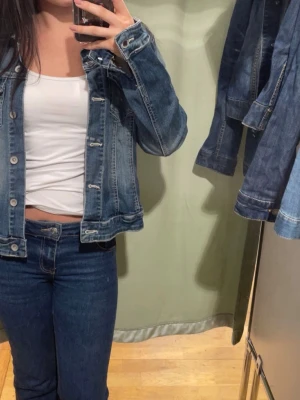 Klassisk blå jeansjacka - Säljer en klassisk blå jeansjacka med raka linjer och silverfärgade knappar. Jackan har två bröstfickor med lock och sömmar i kontrastfärg. Perfekt att slänga över en t-shirt för en avslappnad look.