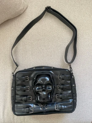 Svart axelväska med spännen och nitar - Cool svart axelväska i glansigt konstläder med flera dekorativa spännen och nitar på framsidan. Väskan har justerbar axelrem och ett unikt mönstrat innerfoder med vita dödskallar. Perfekt för dig som gillar alternativ stil och detaljer. Helt oanvänd, måtten är: Bredd 35 cm, höjd 23 cm, djup 9 cm