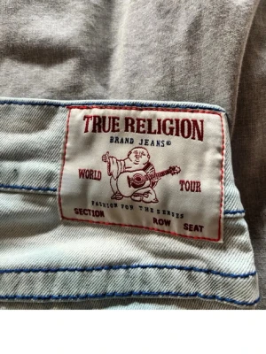 Ljusblå bootcut jeans True Religion - Säljer ett par ljusblå jeans från True Religion med bootcut passform, låg midja och klassiska kontrastsömmar. Jeansen har en ikonisk patch bak med röd text och motiv. Tillverkade i denim, perfekta för en avslappnad och trendig look.