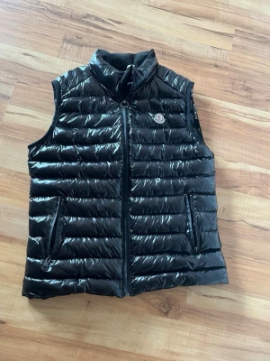 Svart dunväst från Moncler - Snygg svart dunväst från Moncler med glansig finish och ståkrage. Västen har två dragkedjeförsedda fickor fram och Moncler-logga på bröstet. Perfekt att slänga över en hoodie eller tröja för extra värme. Klassisk quiltad design och lätt vaddering.