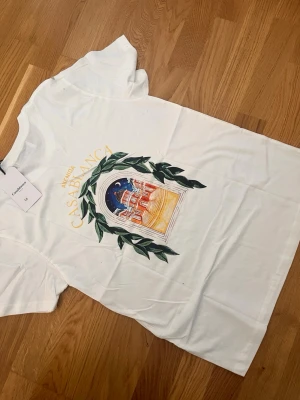 Vit Casablanca t-shirt med tryck - Vit t-shirt från Casablanca med färgglatt tryck framtill som föreställer ett fönster med blad och byggnadsmotiv. Klassisk rund hals och korta ärmar. Tillverkad i mjuk bomull för en skön känsla och stilren look.