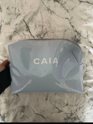 Ljusblå necessär från CAIA - Säljer en ljusblå necessär från CAIA med blank yta och vit logga framtill. Väskan har dragkedja upptill och ett rymligt innerfack med ljuslila foder. Perfekt för smink eller småprylar när du är på språng.