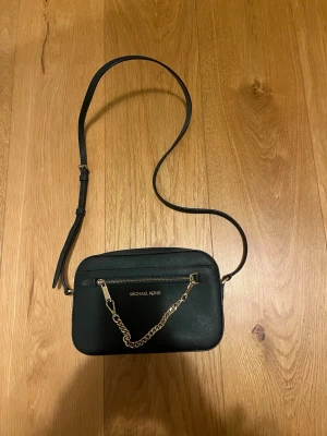 Mörkgrön axelväska Michael Kors - Snygg mörkgrön axelväska från Michael Kors i stilren design. Väskan har guldfärgade detaljer, kedja framtill och justerbar axelrem. Tillverkad i ett tåligt konstläder med dragkedja och logga på framsidan. Perfekt för dig som gillar klassisk och trendig stil.