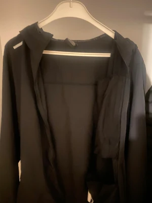 Svart vindjacka från Under Armour - Snygg svart vindjacka från Under Armour med huva och dragkedja framtill. Jackan har vita detaljer och logga på bröstet samt en bröstficka med dragkedja. Tillverkad i lätt och funktionellt material, perfekt för aktiva dagar utomhus.