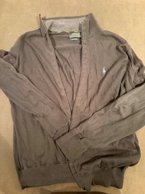 Grå cardigan från Polo Ralph Lauren - Stilren grå cardigan från Polo Ralph Lauren i mjuk pima cotton. Tröjan har dragkedja framtill, ribbade muddar och den klassiska broderade loggan på bröstet. Perfekt för lager på lager och enkel att matcha med olika outfits. Ny pris 2800kr.