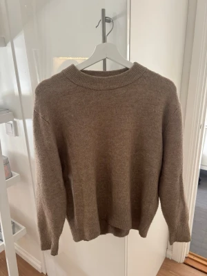 Beige stickad tröja med rund hals - Mysig beige stickad tröja med rund halsringning och ribbade muddar vid ärmslut och nederkant. Tröjan har en avslappnad passform och är perfekt för lager på lager. Materialet ser ut att vara mjukt och värmande, troligen ullblandning.