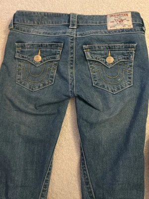 Blå True Religion jeans med fickdetaljer - Snygga blå jeans från True Religion med ikoniska kontrastsömmar och dekorativa ficklock bak. Klassisk femficksmodell med guldfärgade knappar och detaljer. Tillverkade i mjuk denim som sitter skönt och har en tidlös look.