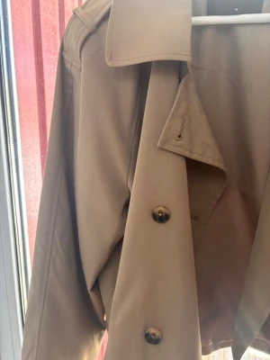 Kort beige trenchcoat från ONLY - Snygg kort trenchcoat i beige från ONLY med klassisk krage och stora knappar framtill. Jackan har axelklaffar och raka ärmar, perfekt för en stilren look. Materialet är slätt och ger en clean känsla. Passar dig som gillar minimalistisk och tidlös stil.