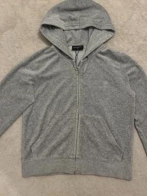 Grå hoodie från Juicy Couture - Snygg grå zip-hoodie från Juicy Couture med huva och dragsko. Mjuk bomullsblandning, två fickor fram och JC-logga broderad på bröstet. Perfekt för en chill och avslappnad stil.