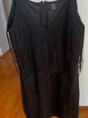 Svart fransklänning från SHEIN - Cool svart kortklänning från SHEIN med långa fransar framtill och baktill. Klänningen är ärmlös, har v-ringning och dragkedja i ryggen. Materialet är troligtvis polyester eller syntet, vilket ger en mjuk och följsam känsla. Perfekt för dig som vill sticka ut med en unik look.