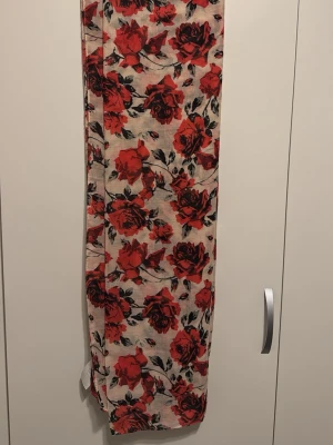 Beige sjal med röda rosor - Snygg beige sjal med stora röda rosor och svarta blad. Tunn och luftig, perfekt att slänga över axlarna för en cool och romantisk look. Mönstret är riktigt iögonfallande och ger en trendig vibe till vilken outfit som helst.