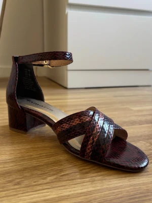 Rödbruna sandaler med ormmönster Tamaris - Säljer ett par rödbruna/burgundy sandaler från Tamaris med snyggt ormmönster och bred klack. Sandalerna har remmar över foten och runt ankeln med guldfärgat spänne. Materialet är konstläder och innersulan har Touch it-funktion för extra komfort.