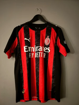 AC Milan fotbollströja Puma röd/svart - AC Milan matchtröja från Puma med klassiska röda och svarta vertikala ränder, klubbmärke på bröstet och vit Puma-logga. Tillverkad i lätt polyester med tryckta sponsorloggor och detaljer. Perfekt för dig som älskar fotboll och vill representera Milan. Storlek S.