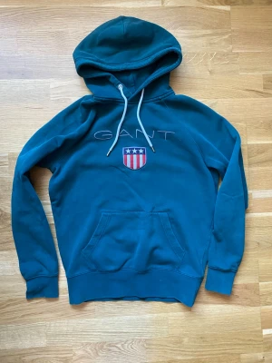 Gant hoodie - Snygg hoodie från GANT med huva och vita dragsnören. På bröstet finns GANTs broderade logga och ett sköldmärke med stjärnor och ränder. Hoodien har en klassisk känguruficka och ribbade muddar. Perfekt för en avslappnad och sportig stil. Kan gå ner i pris vid snabb affär.