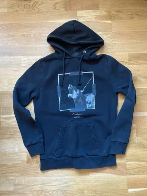 Svart hoodie - Svart hoodie från med tryck av en  skällande hund  i vitt och grått på bröstet. Klassisk känguruficka framtill, ribbade muddar och dragsko i huvan. Tillverkad i mjukt bomullsmaterial för skön känsla. Perfekt hoddie .Kan gå ner i pris vid snabb affär.