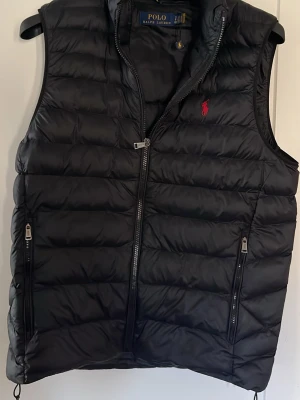 Svart dunväst från Polo Ralph Lauren - Snygg svart dunväst från Polo Ralph Lauren med quiltad design och två dragkedjefickor framtill. Västen har hög krage och den klassiska röda logotypen broderad på bröstet. Perfekt för lager-på-lager och håller dig varm med stil. Ny pris 3600kr köpt på zalando mitt pris 2800kr västen är i ny skick utan skador.