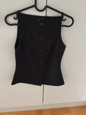 Svart topp med knappar - Stilren svart topp med fem knappar framtill och hög halsringning. Toppen har en figurnära passform och är ärmlös, vilket ger en clean och modern look. Tillverkad i ett slätt material som känns bekvämt och passar till många olika outfits.