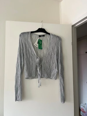 Ljusgrå stickad cardigan med knyt - Trendig ljusgrå stickad cardigan från Bik Bok med vågigt hålmönster och knyt framtill. Croppad modell med långa ärmar och djup V-ringning. Perfekt att slänga över ett linne eller klänning för en cool lager-på-lager-look.
