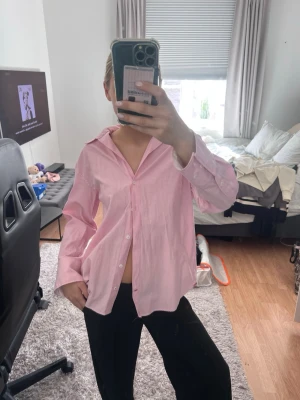 Ljusrosa randig skjorta - Säljer en ljusrosa skjorta med tunna vita ränder. Skjortan har klassisk krage, knäppning framtill och långa ärmar med manschetter. Passformen är loose och materialet känns som bomull, perfekt för en avslappnad stil.