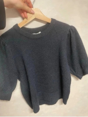 Mörkgrå stickad tröja med puffärm - Mysig mörkgrå stickad tröja med korta puffärmar och rund halsringning. Tröjan har ribbstickade muddar vid ärmslut och nederkant, vilket ger en snygg och trendig look. Perfekt att styla med jeans eller kjol. Aldrig använd