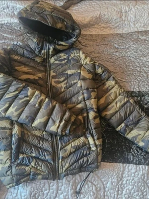Kamo-mönstrad dunjacka med huva - Säljer en dunjacka med snyggt kamouflagemönster i gröna och beige toner. Jackan har huva, dragkedja framtill och två sidofickor. Perfekt för kalla vinterdagar och riktigt skön att bära. Passar dig som gillar streetwear och vill sticka ut.