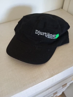 Svart keps Djurtjänst Bollnäs - Snygg svart keps med böjd skärm och broderad text 'Djurtjänst i Bollnäs AB' framtill. Klassisk modell i bomullstyg med justerbar baksida. Perfekt för dig som vill ha en enkel och stilren keps med unik logga.