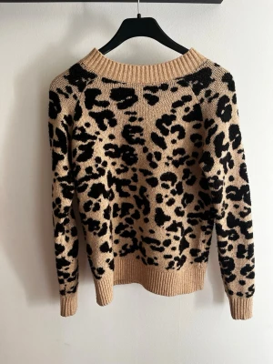 Beige leopardmönstrad stickad tröja - Stickad tröja från Vero Moda i beige med svart leopardmönster. Tröjan har rund halsringning, ribbade muddar och är tillverkad i ett mjukt material som passar perfekt för kyligare dagar. Snygg och trendig design som sticker ut. Superfin men börjar tyvärr bli för tajt för mig.