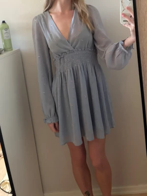 Ljusblå kortklänning med v-ringning - Söt ljusblå kortklänning med v-ringad omlottdesign och långa, transparenta ärmar. Klänningen har smock i midjan för en bekväm passform och små diskreta prickar i tyget. Perfekt för sommarens alla tillfällen.