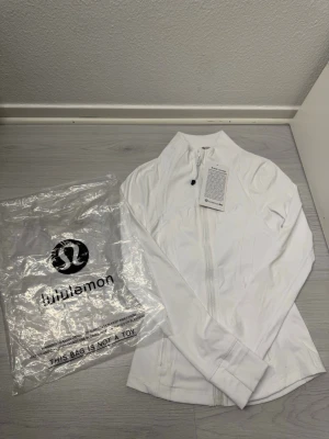 Vit zip-kofta från Lululemon - Slimmad vit träningsjacka från Lululemon med hel dragkedja framtill, hög krage och två fickor med dragkedja. Tillverkad i ett stretchigt funktionsmaterial som passar perfekt till träning eller som ett extra lager. Klassisk och stilren design. Är ej använd taggen sitter fortfarande kvar påse följer med. Den ser liten ut på bilden men sitta ganska bra i när man har på sig den. Fraktar samma dag eller dagen efter. Storlek S/XS. DM för fler frågor :)